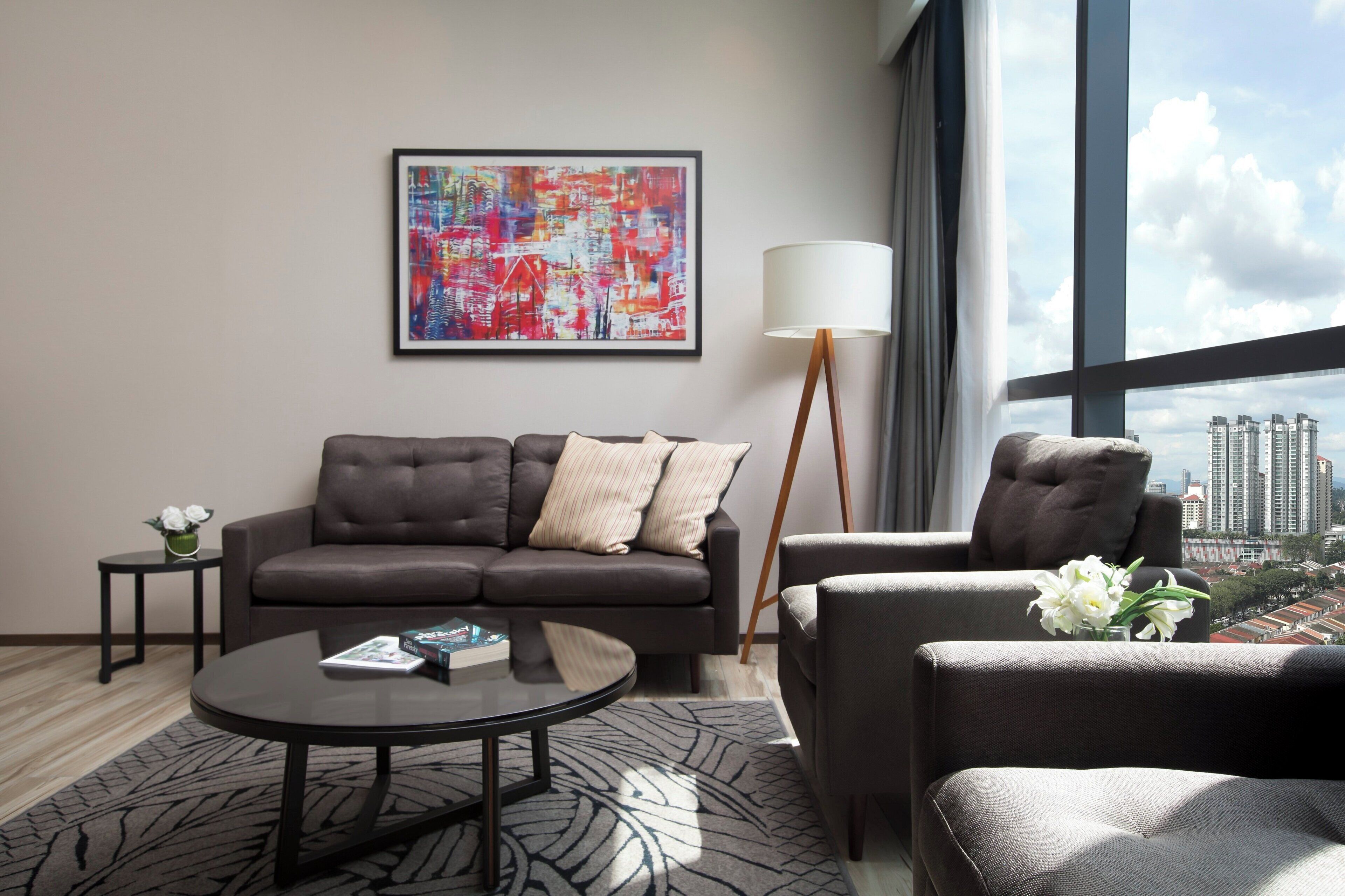 A modern living room with a grey sofa, a round coffee table, and a floor lamp. A colorful abstract painting is on the wall, and a cityscape is visible through the window.