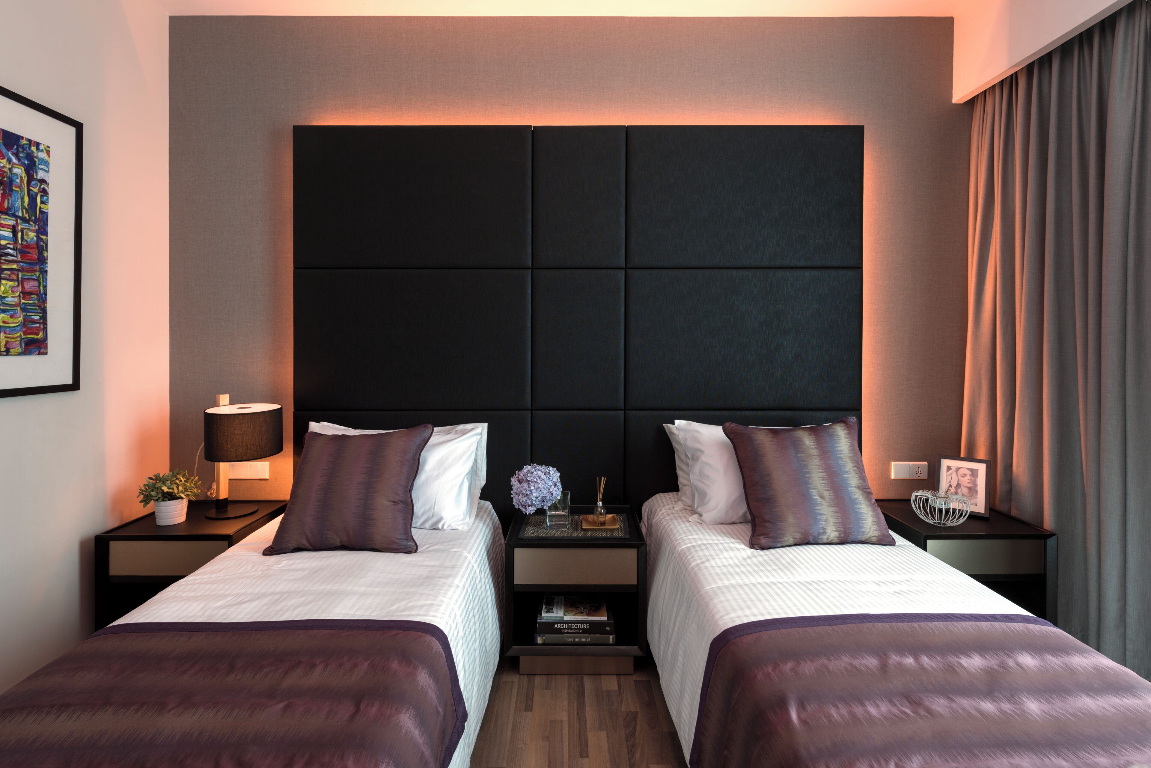 A modern hotel room with two beds, a large padded headboard, bedside tables with lamps, and a framed picture.