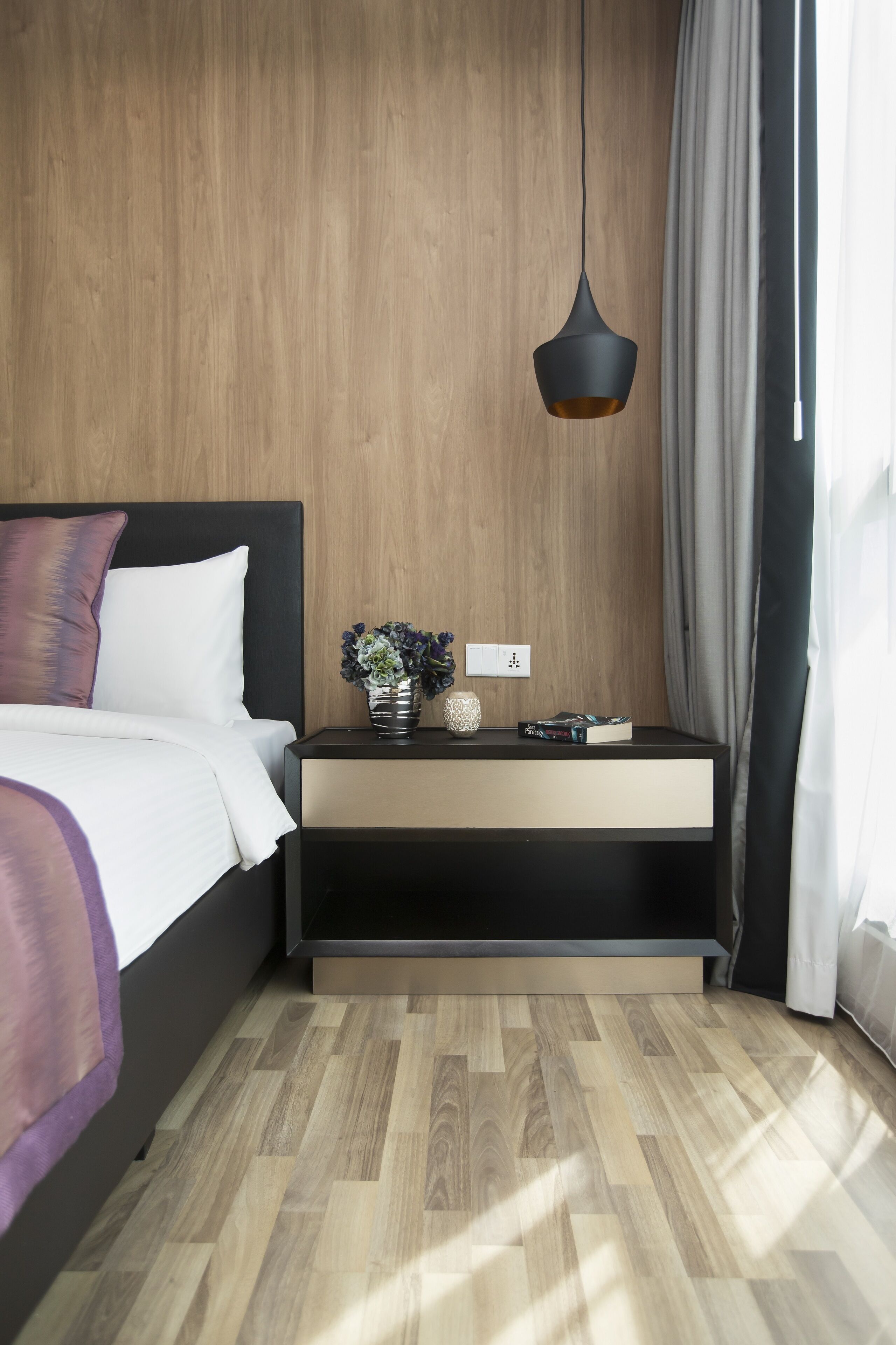A modern bedroom with a wooden wall, a bed with white bedding, a bedside table with a vase of flowers, and a hanging pendant light.