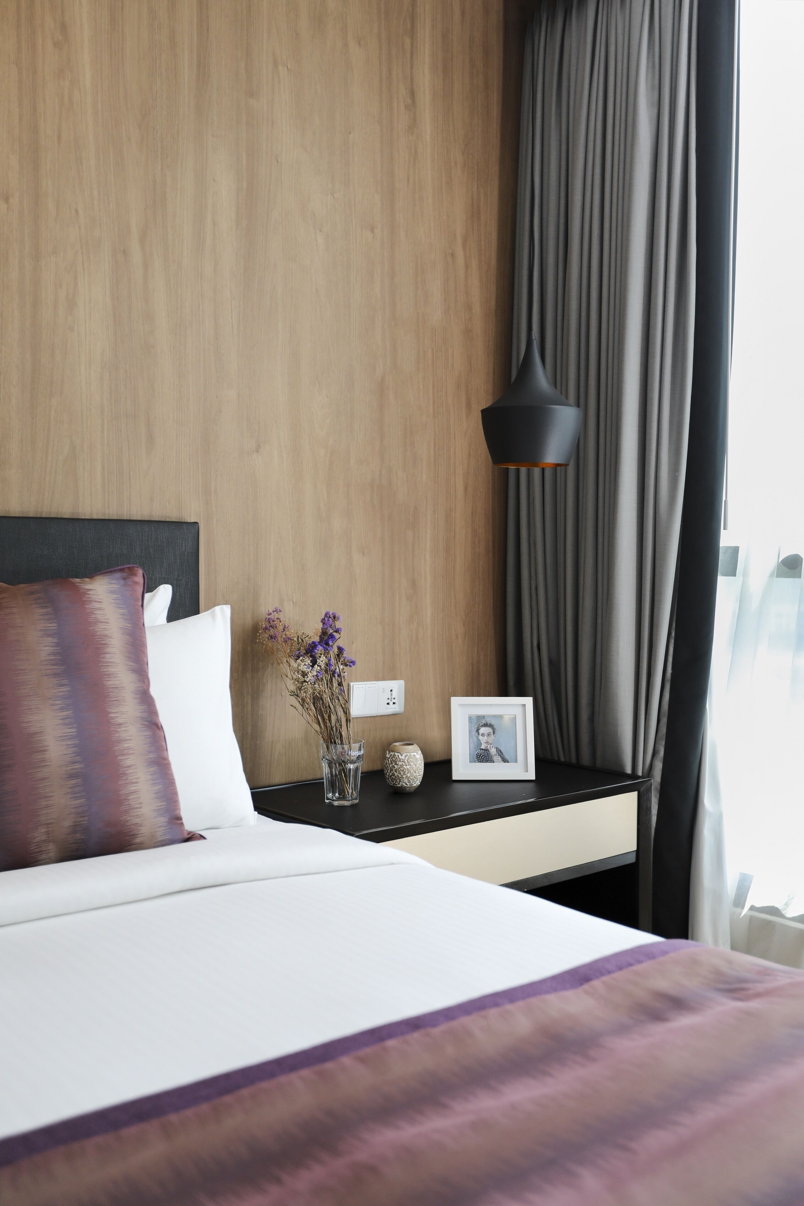 A neatly made bed with a white and purple blanket, a dark headboard, and a bedside table with a vase of dried flowers, a small vase, and a framed photo.