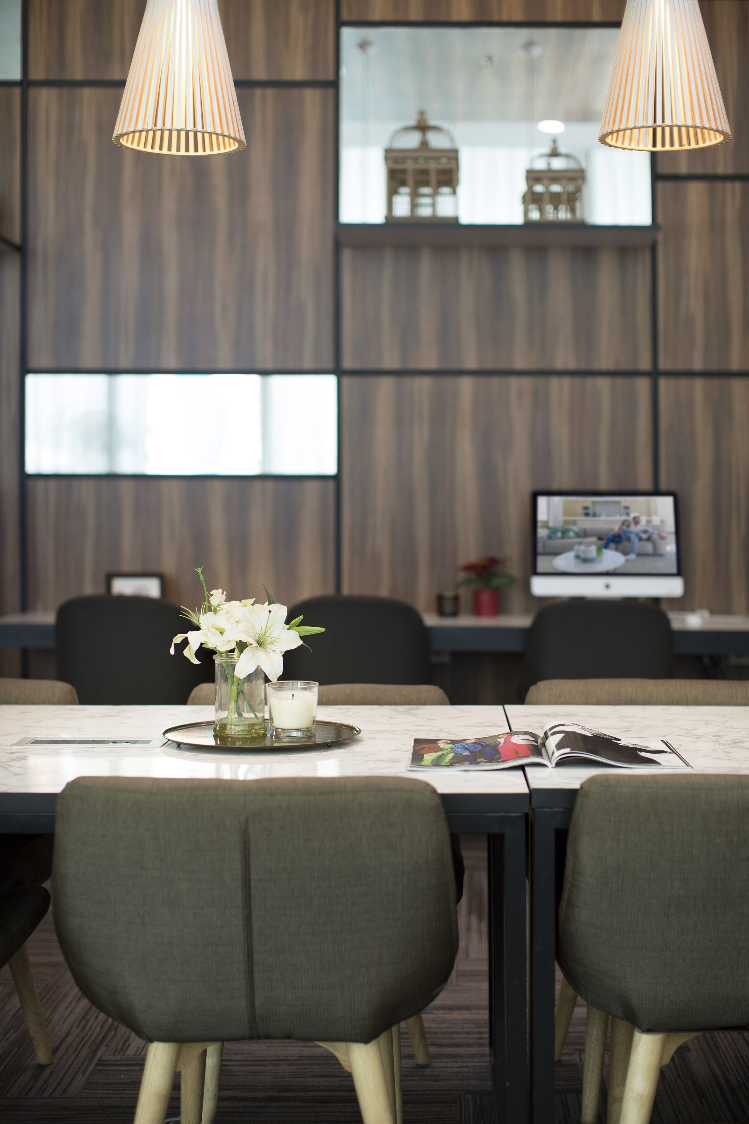 A modern dining area with a wooden wall, two pendant lights, a vase with white flowers, and a table with magazines.