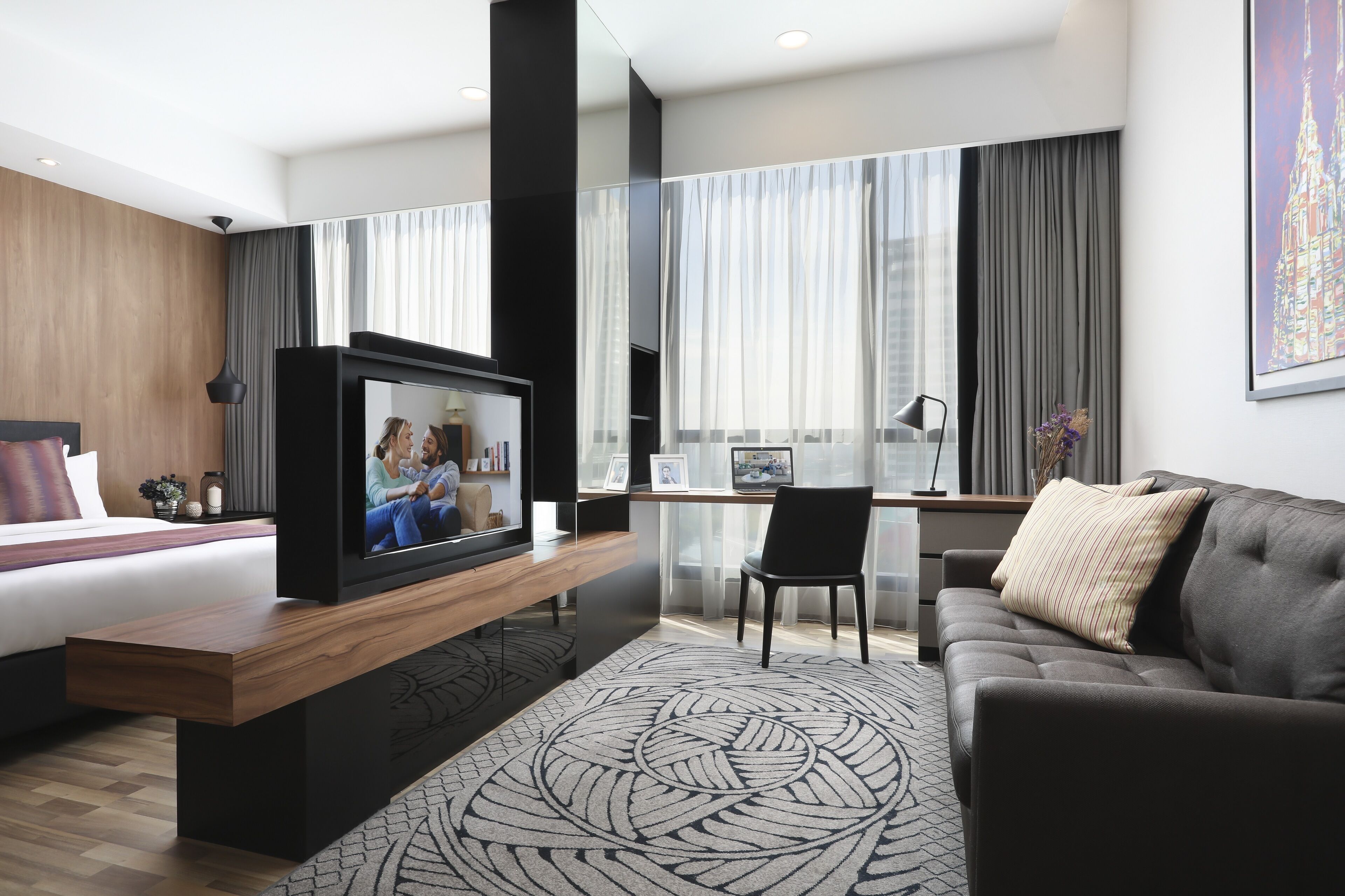 A modern hotel room with a bed, a TV on a wooden stand, a desk with a computer, a grey sofa, and a large window with curtains.