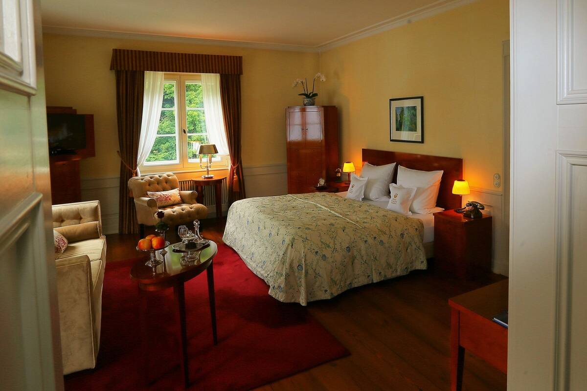 Een hotelkamer met een bed, een zithoek met een bank en fauteuils, een eettafel met een bloevas en een televisie.