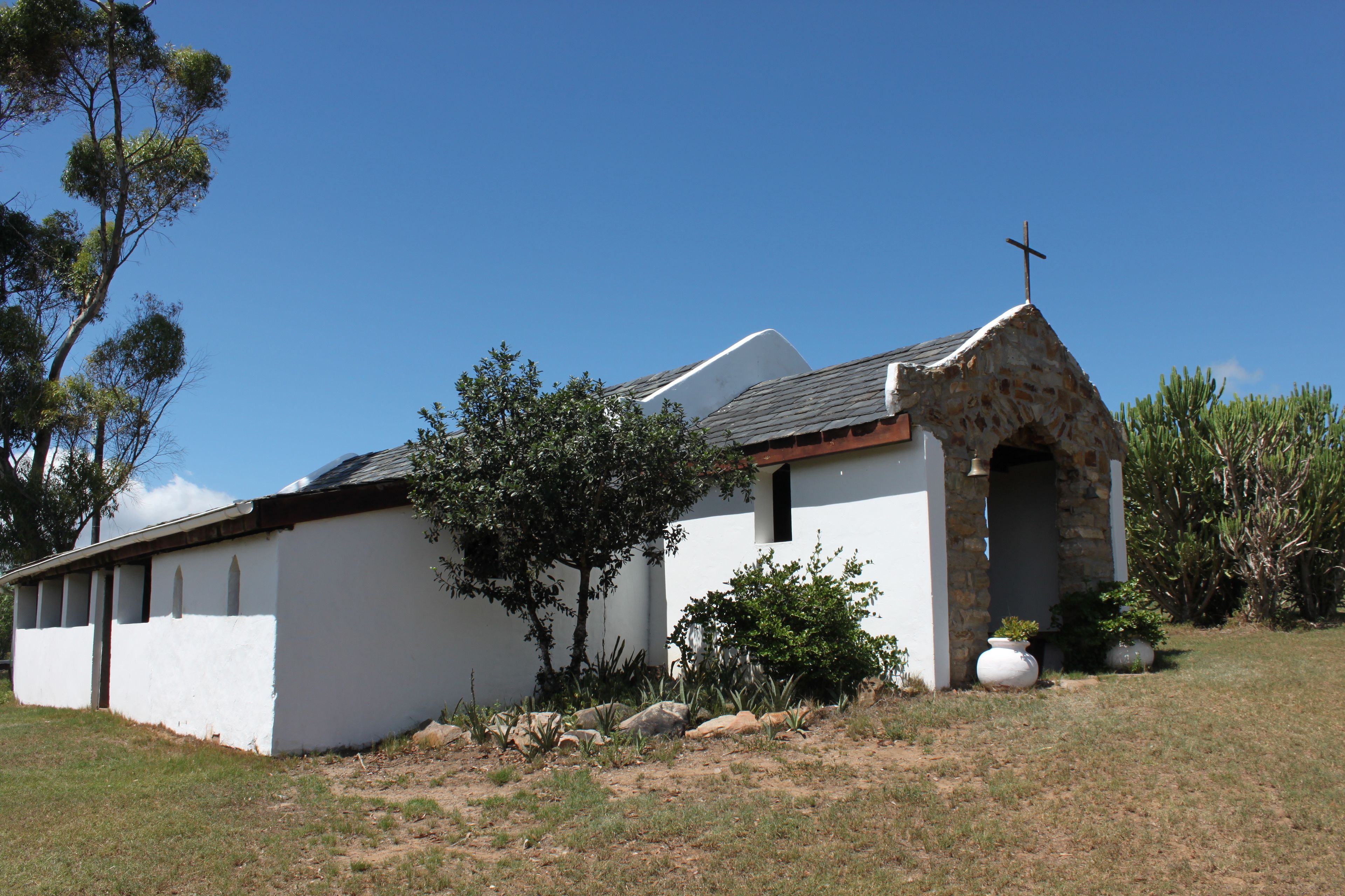 Une petite église blanche, surmontée d’une croix, entourée d’arbres et de plantes.