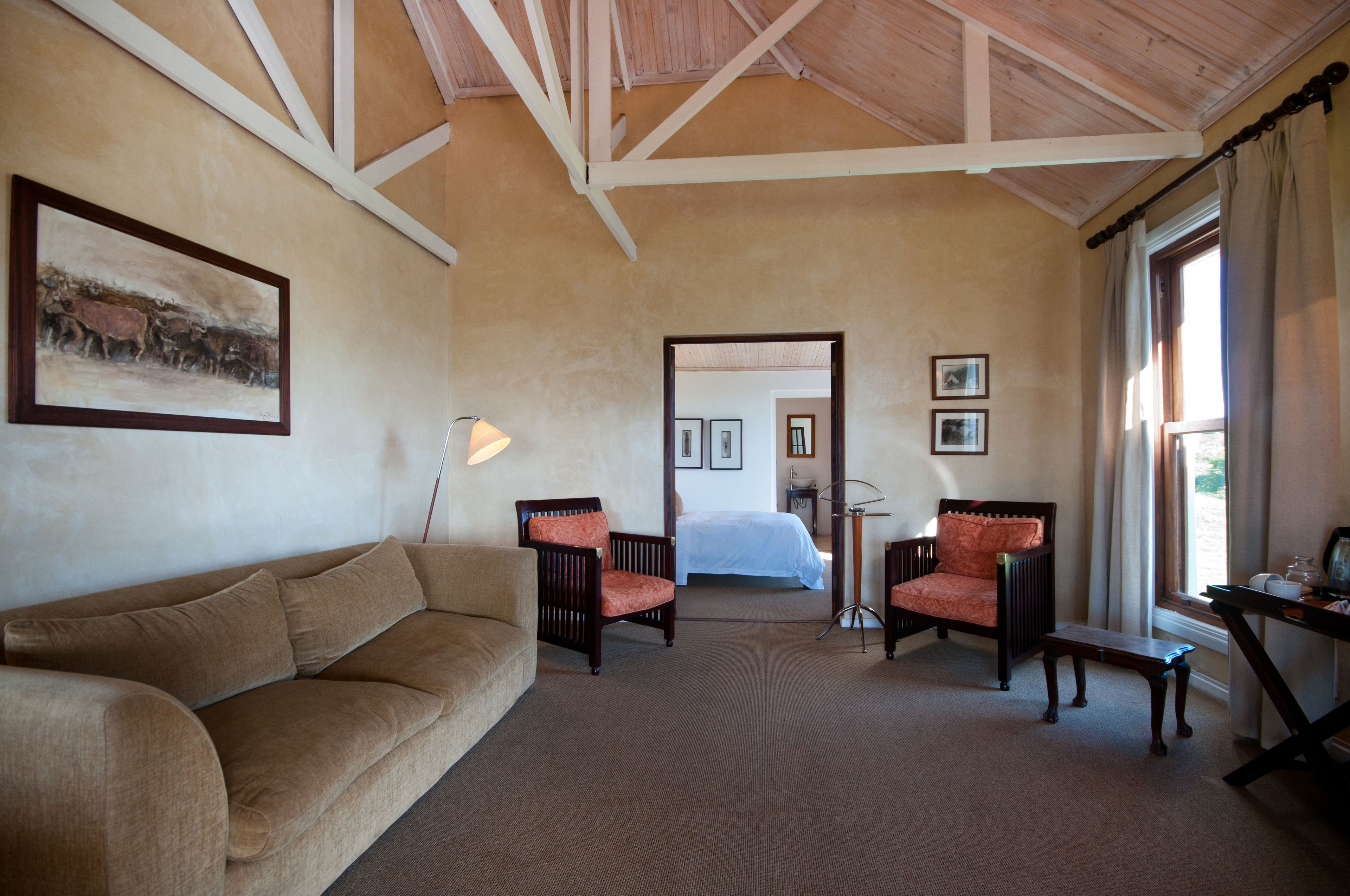 A room with a beige sofa, two armchairs, a small table, and a lamp. A bed is visible in the background.