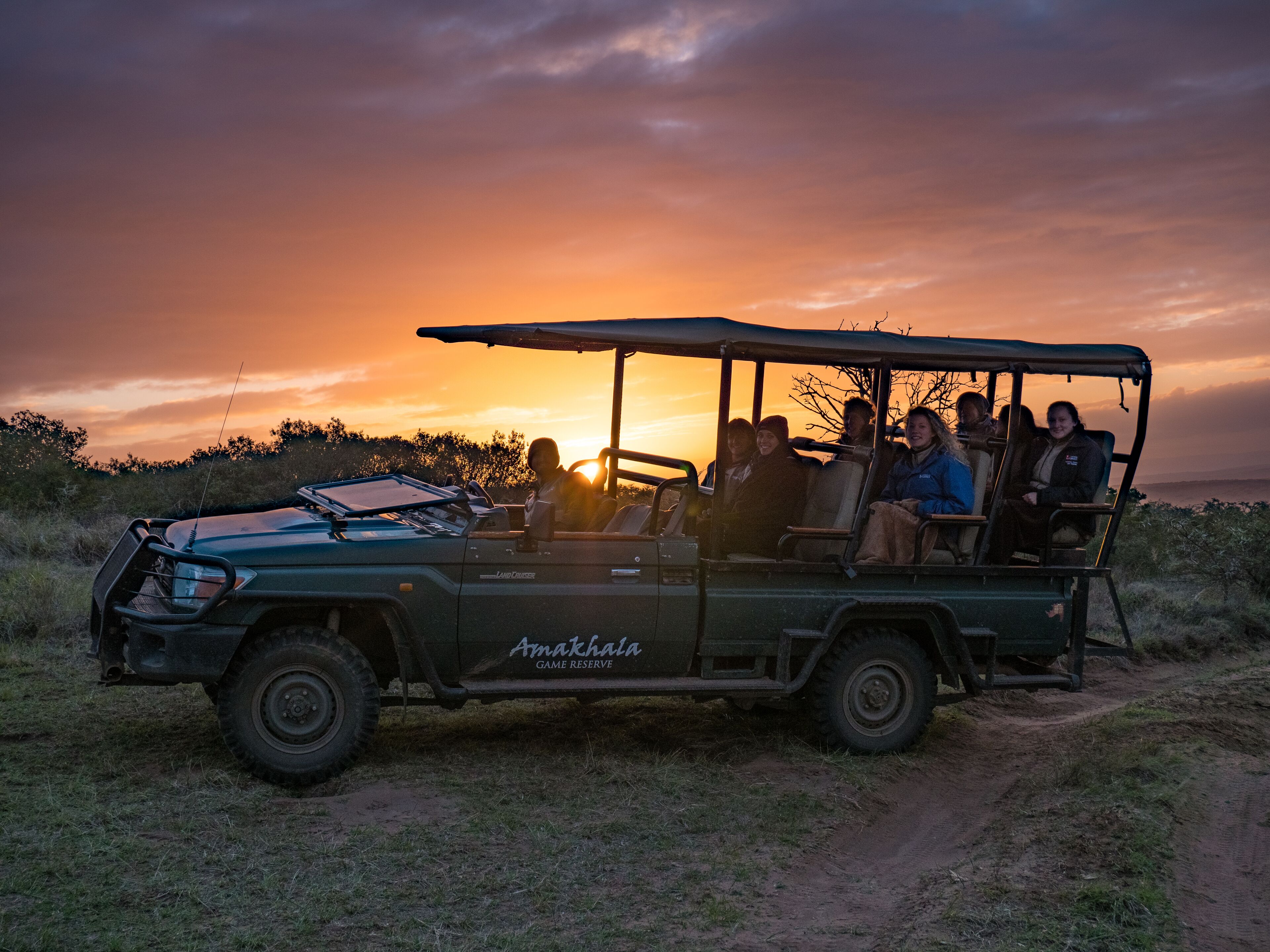 A safari vehicle with passengers, parked on a dirt path with a sunset in the background.