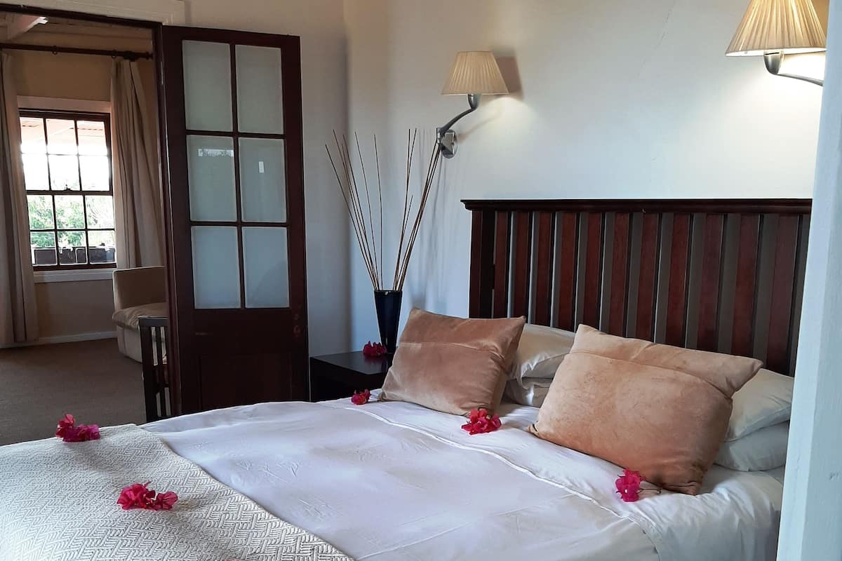A neatly made bed with a patterned bedspread, two pillows, and rose petals. There is a bedside lamp, a vase with reed sticks, and a window with curtains.