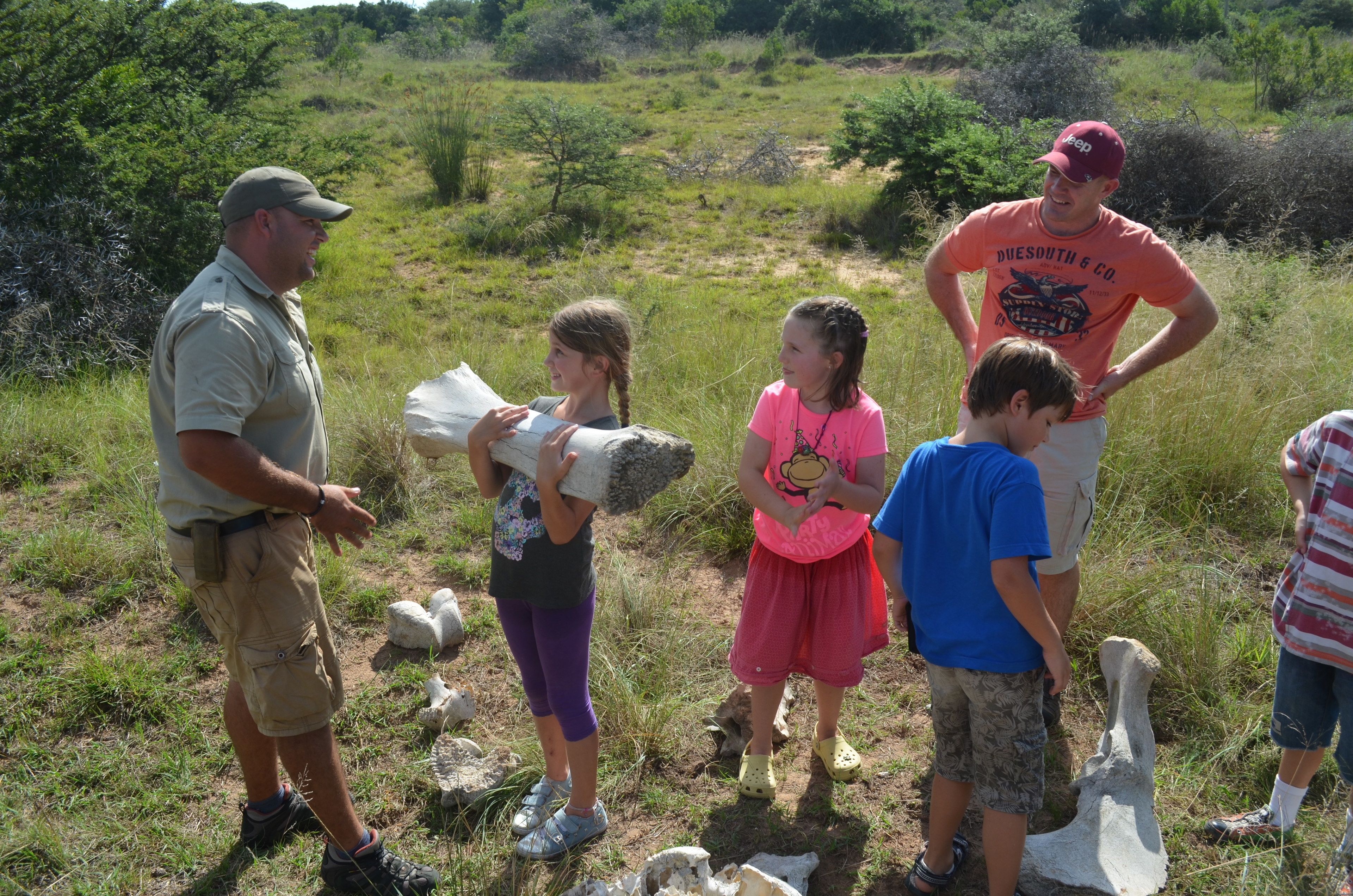 A group of people outdoors in a grassy area with scattered rocks and bushes.