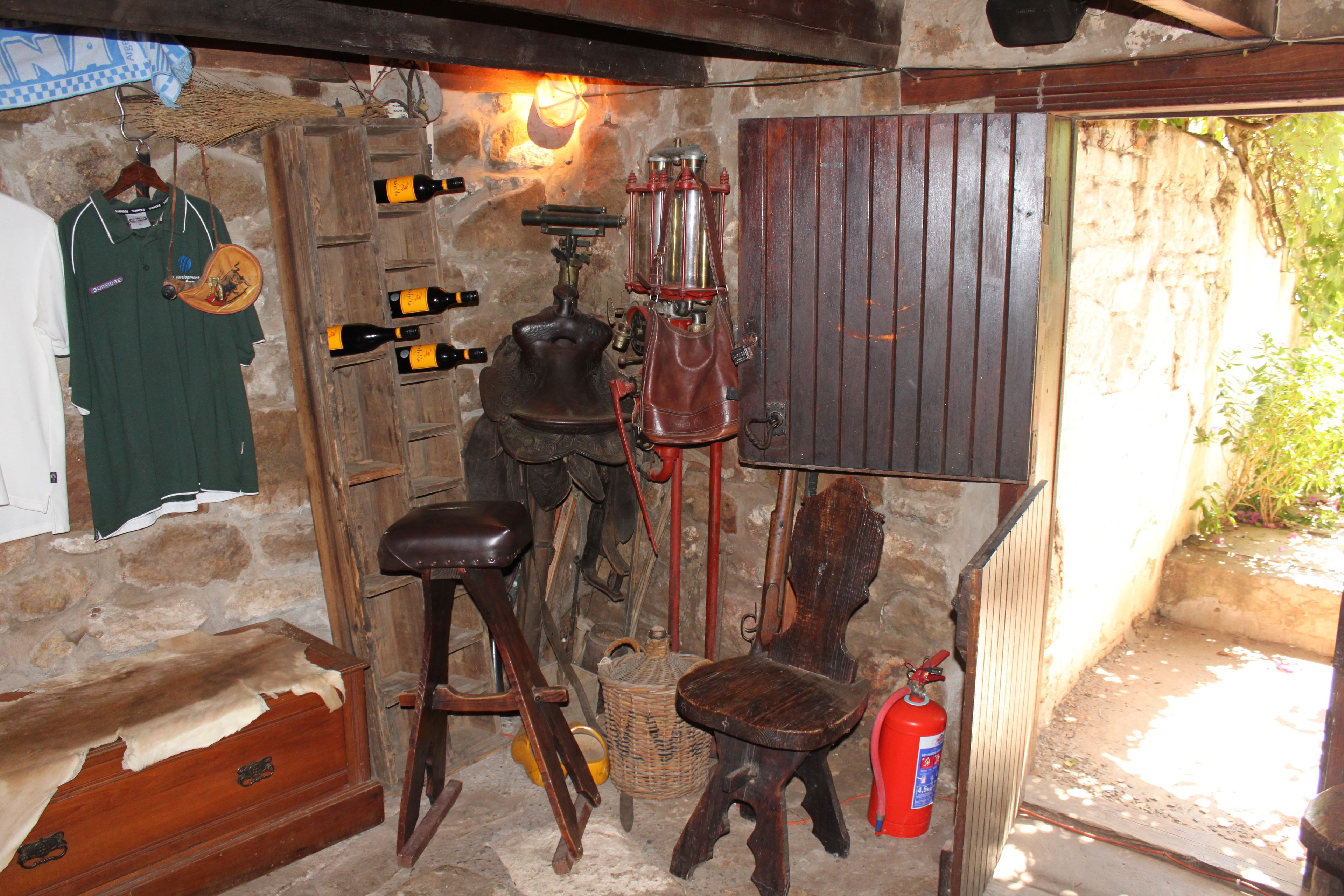 Quarto rústico com um banco de madeira, uma cadeira, um extintor de incêndio e uma prateleira na parede com garrafas e uma camisa verde.
