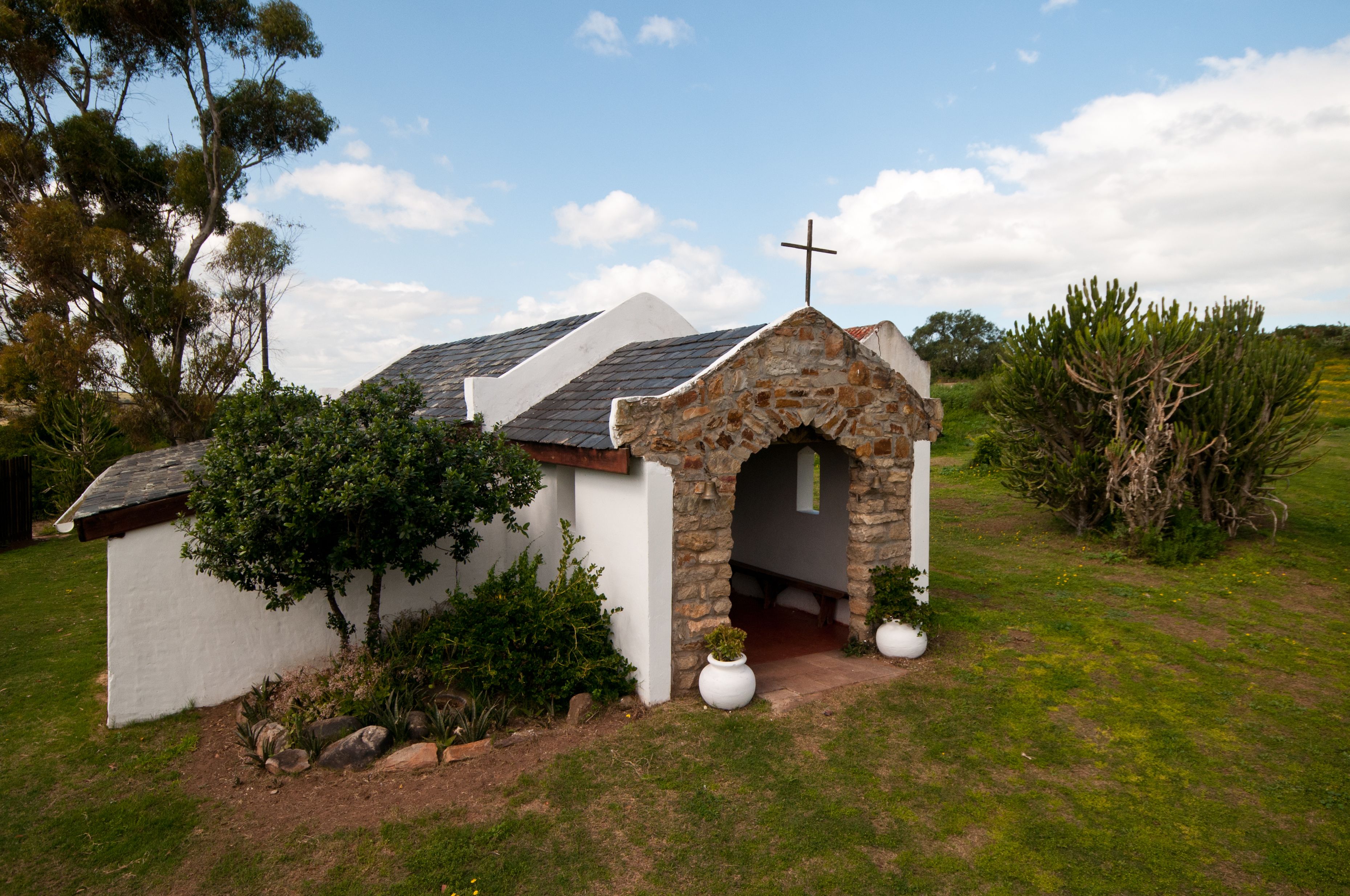 A small stone chapel with a cross, surrounded by greenery and a white building.