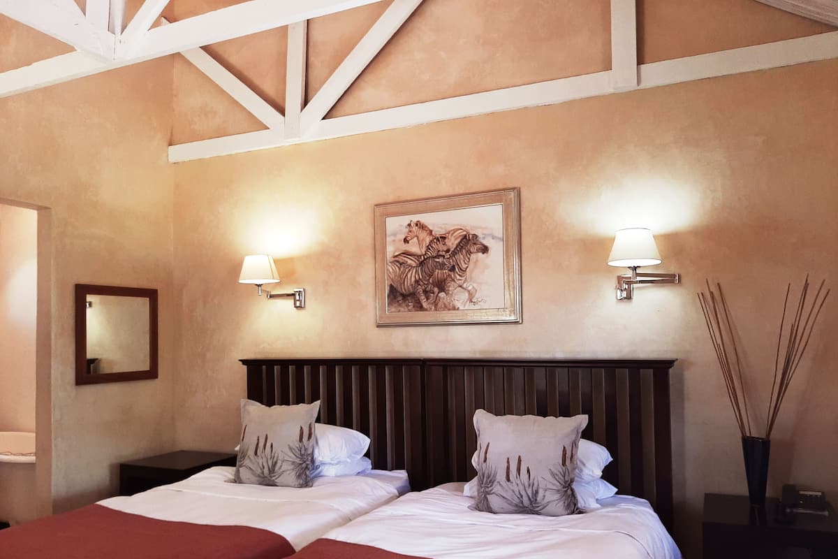 A hotel room with two beds, a wooden headboard, a framed zebra painting, wall-mounted lamps, and a mirror.