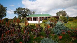 A house with a green roof and white exterior, surrounded by a garden with various plants and trees. - Leeuwenbosch Shearers Lodge (Paterson)