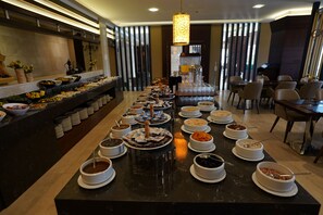 A buffet setup with various food items and beverages arranged on a long table, surrounded by chairs and a modern interior design. - Safir Hotel (Gaziantep)