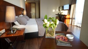 Una habitación de hotel con una cama bien hecha, una mesita de noche con una lámpara, un jarrón con flores, un menú del restaurante y un televisor al fondo.