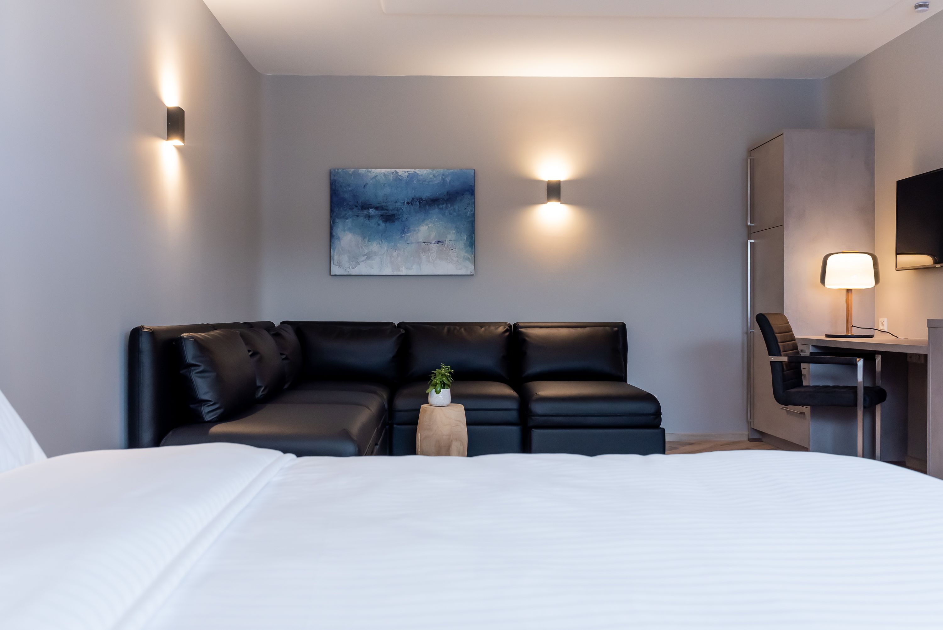 A modern hotel room with a large bed, a black leather sofa, a small wooden side table with a plant, and a desk with a lamp.