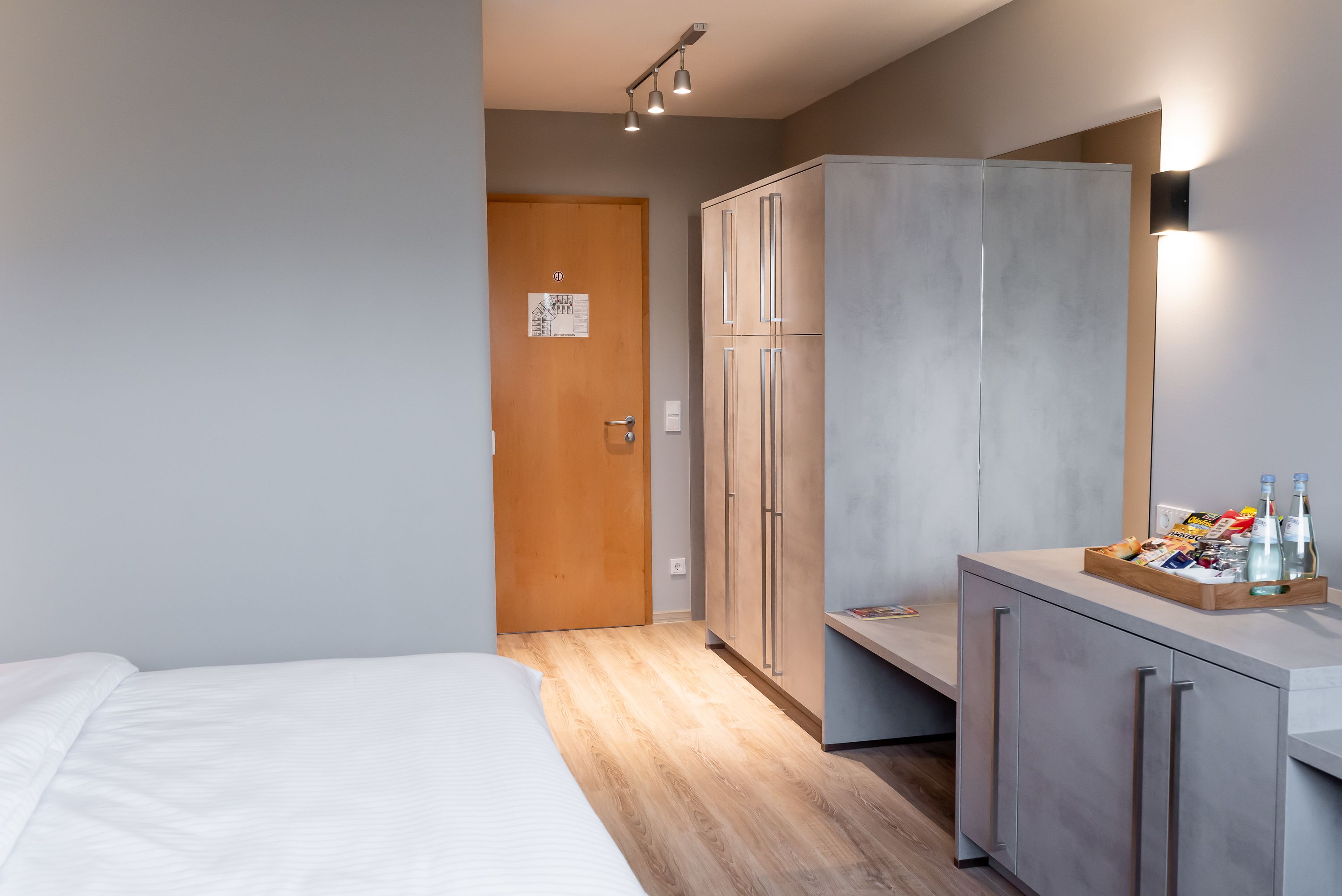 A modern hotel room with a bed, wardrobe, and a small kitchenette area with a tray of snacks and drinks.