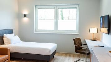 Een moderne hotelkamer met een bed, een bureau, een stoel en een raam met uitzicht op bomen.