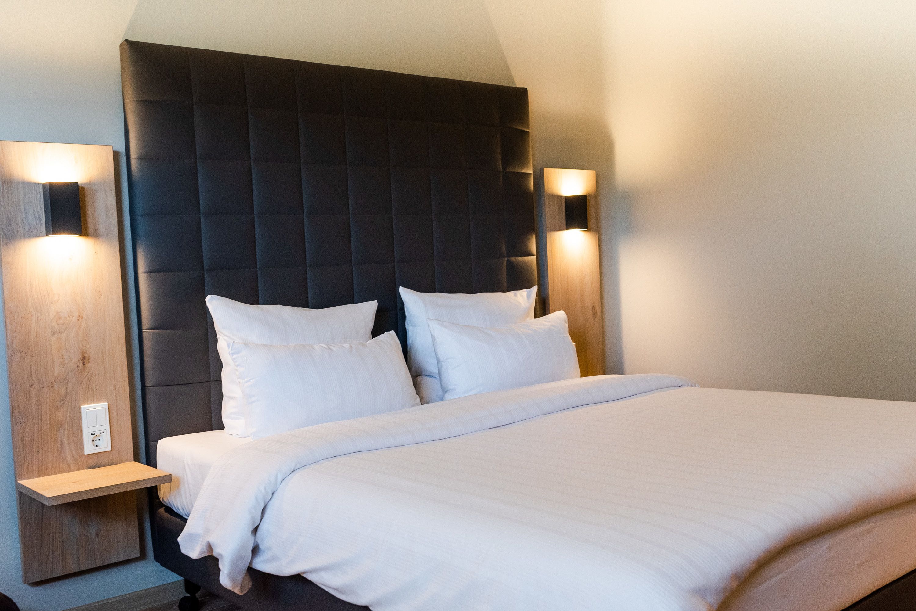 Una cama bien tendida con sábanas blancas y un cabecero oscuro, con lámparas fijadas a la pared a cada lado.