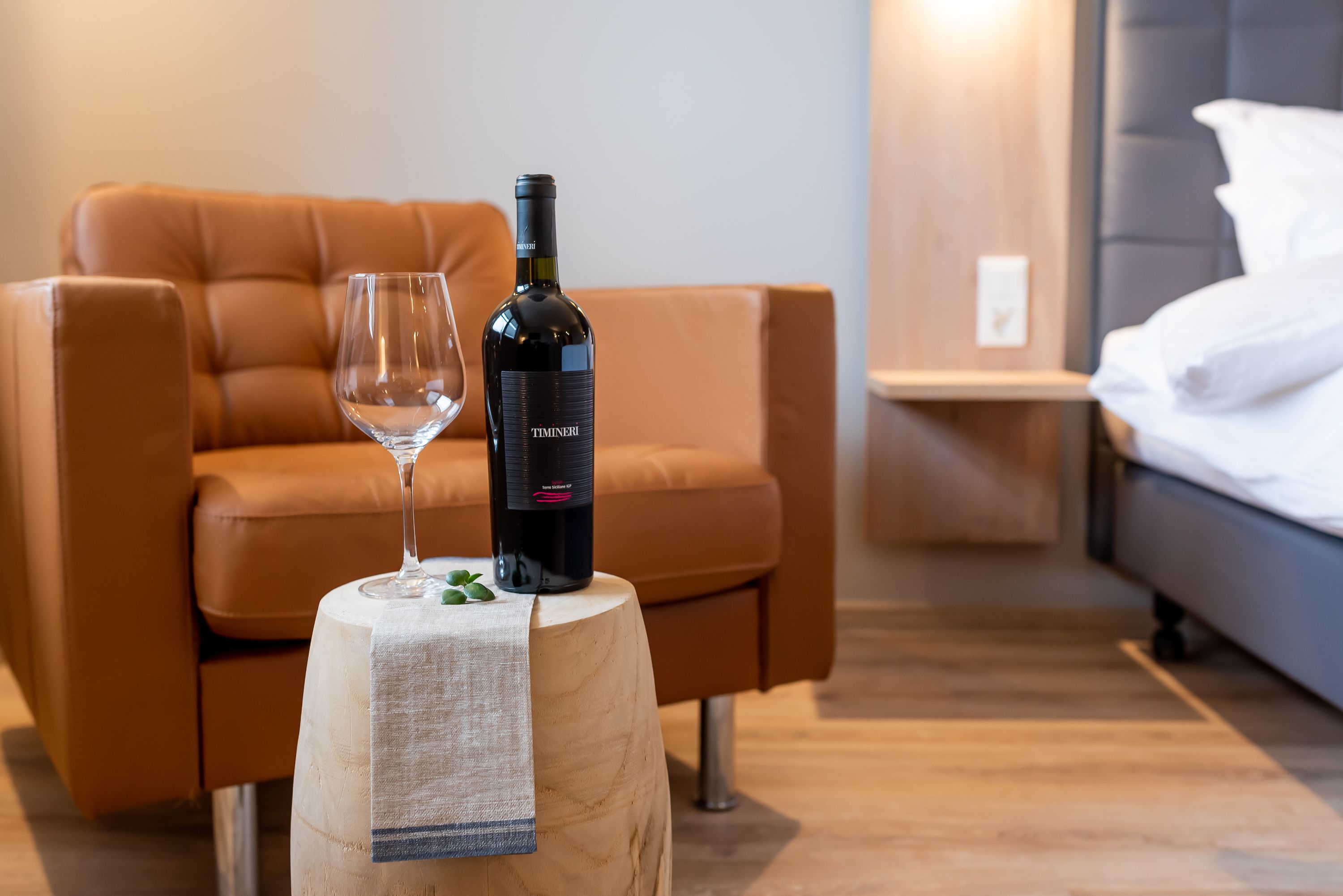 A modern hotel room with a wooden nightstand, a bottle of wine, and a glass on it.