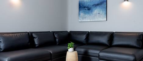 Una sala de estar moderna con un sofá seccional de cuero negro, una mesita de madera con una planta y un cuadro abstracto azul en la pared.
