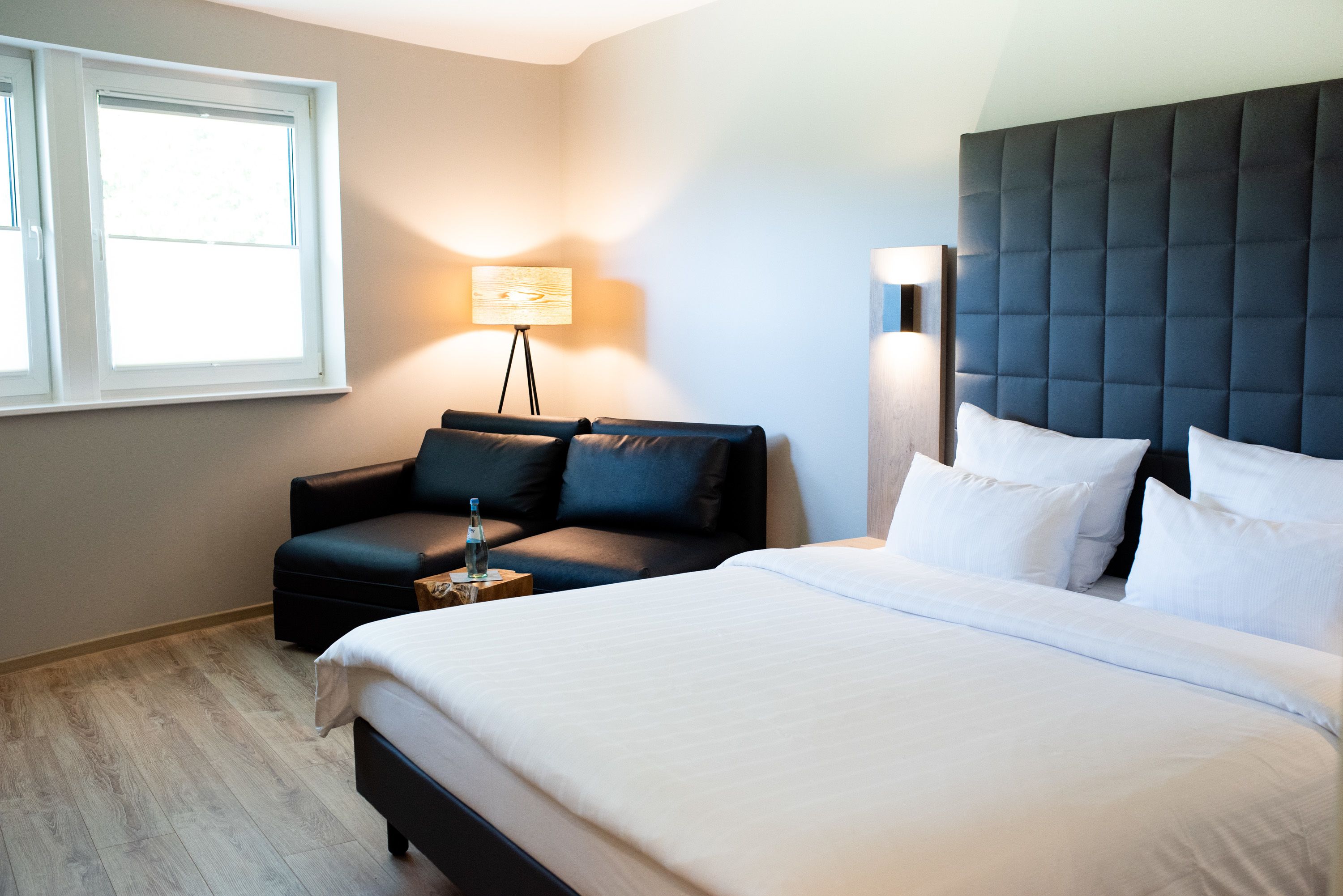 A modern hotel room with a large bed, a sofa, a standing lamp, and a window.