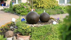 Un jardín con dos esculturas de piedra esféricas, diversas plantas en macetas y un sendero de ladrillo.