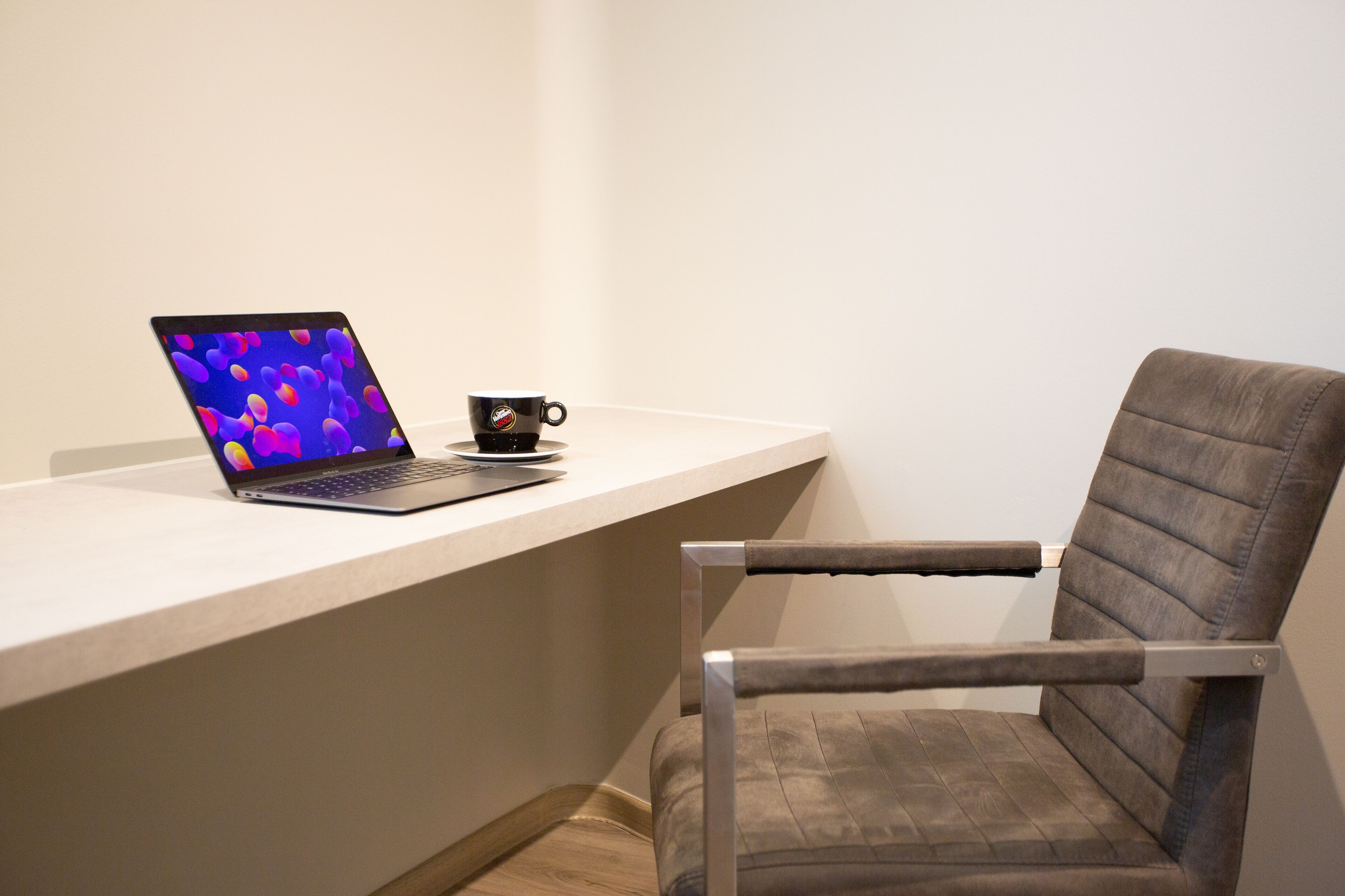 Ein moderner Arbeitsplatz mit einem Laptop auf einem weißen Schreibtisch, einer schwarzen Tasse und einem grauen Stuhl.