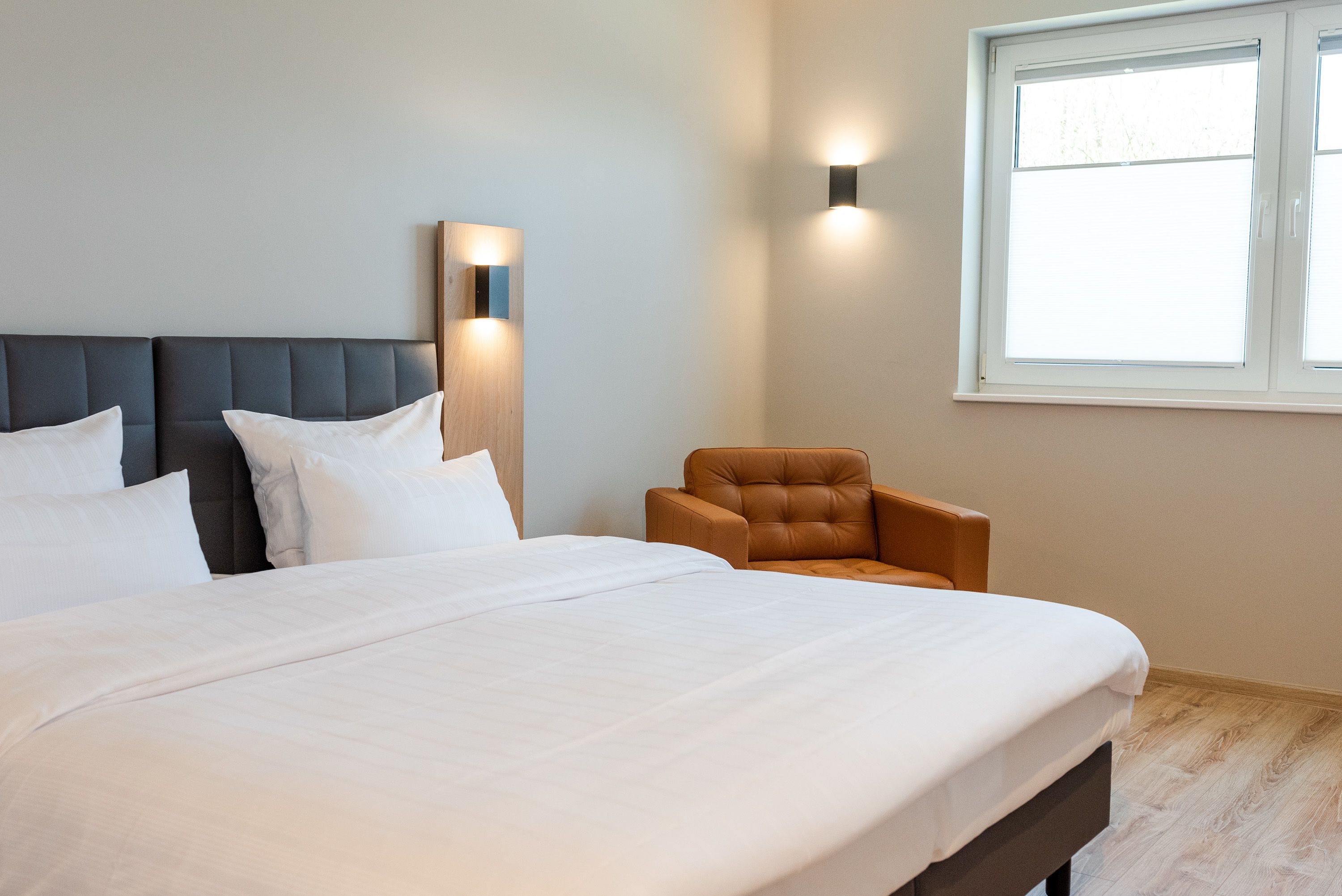 A neatly made bed with white linens, a dark headboard, and a brown armchair beside it.