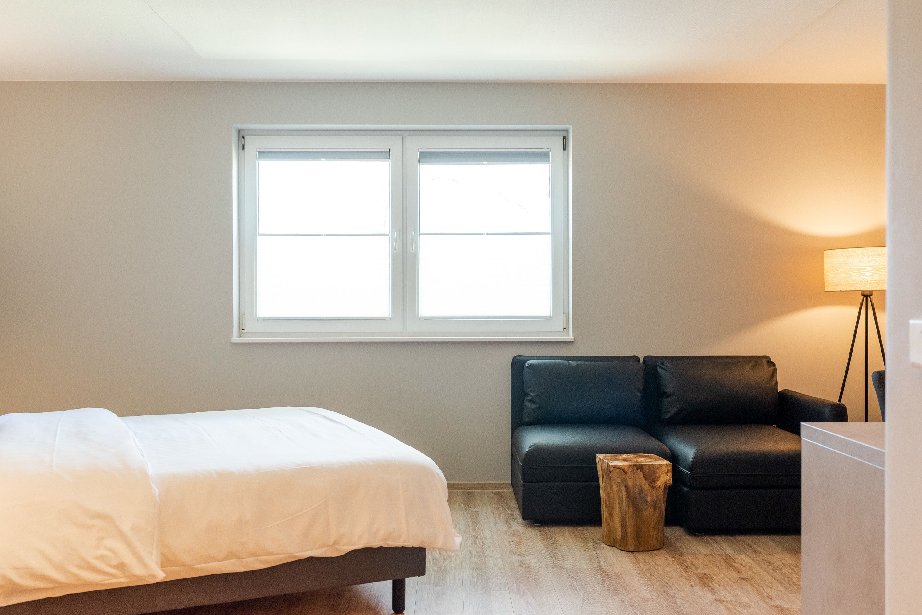 Ein Schlafzimmer mit einem Bett, einer schwarzen Ledersofa, einem Holzschemel und einem Fenster mit Jalousien.