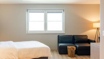 Ein Schlafzimmer mit einem Bett, einer schwarzen Ledersofa, einem Holzschemel und einem Fenster mit Jalousien.