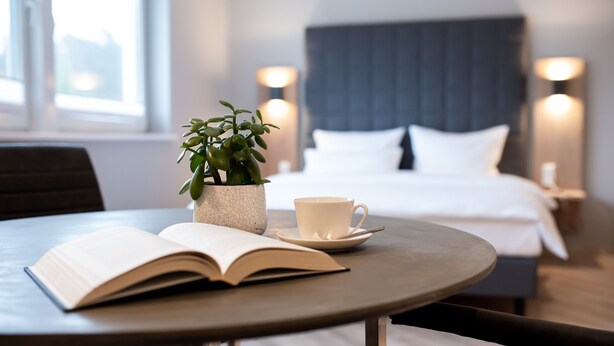 Habitación de hotel con cama, lámparas de noche, una planta en maceta, una taza y un libro abierto sobre una mesa redonda.