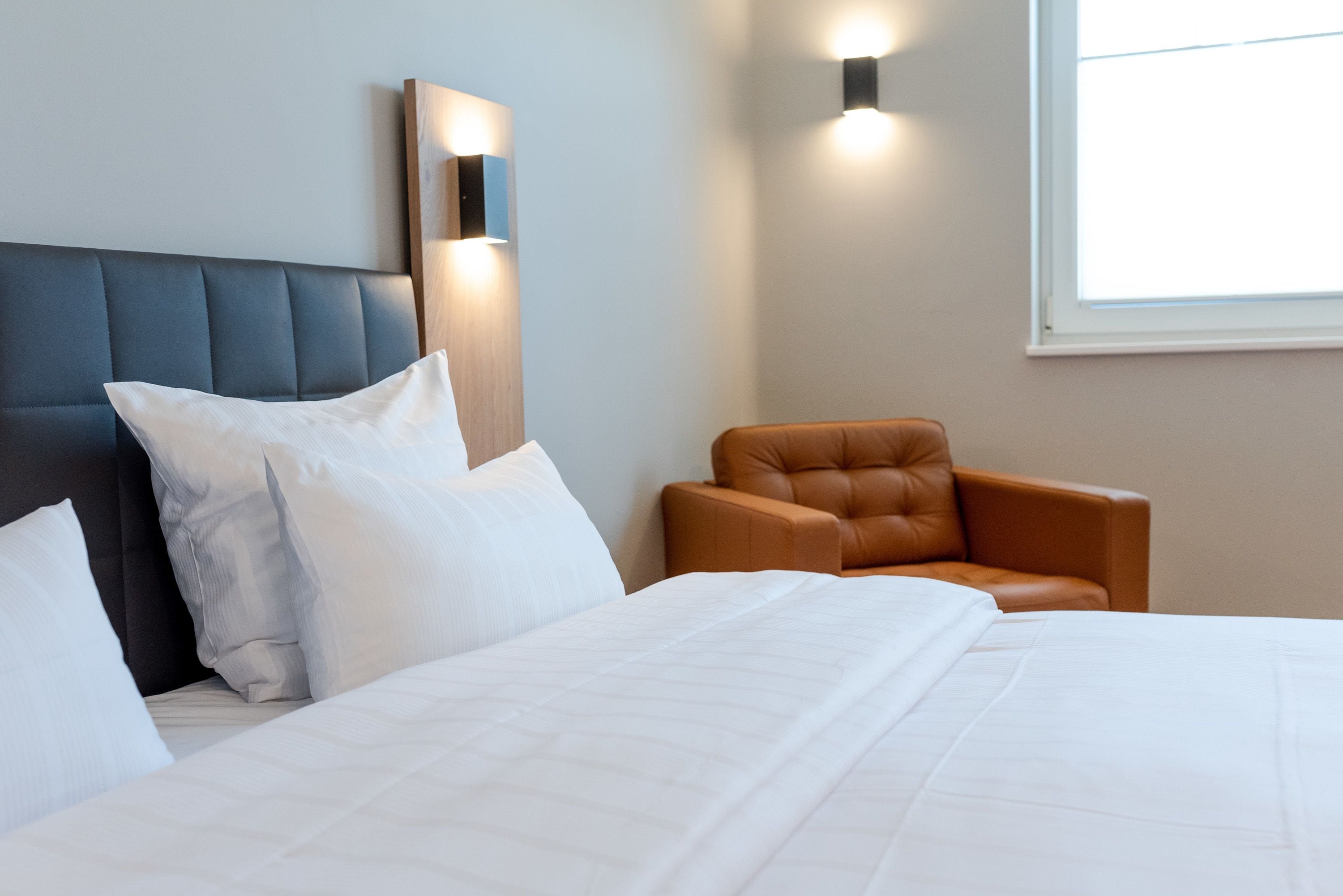 A hotel room with a large bed, a headboard, a brown leather armchair, and wall-mounted lighting.