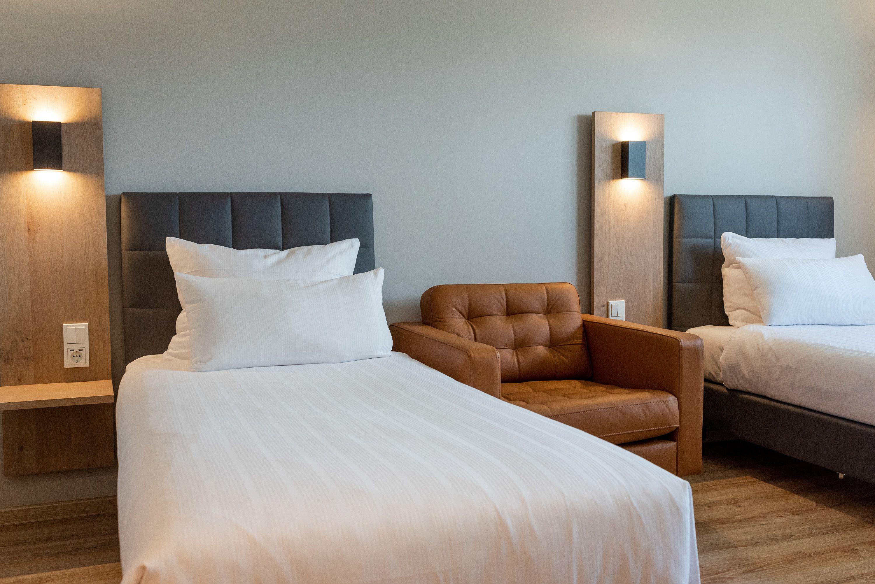 A hotel room with two beds, a brown leather armchair, and wall-mounted lights.