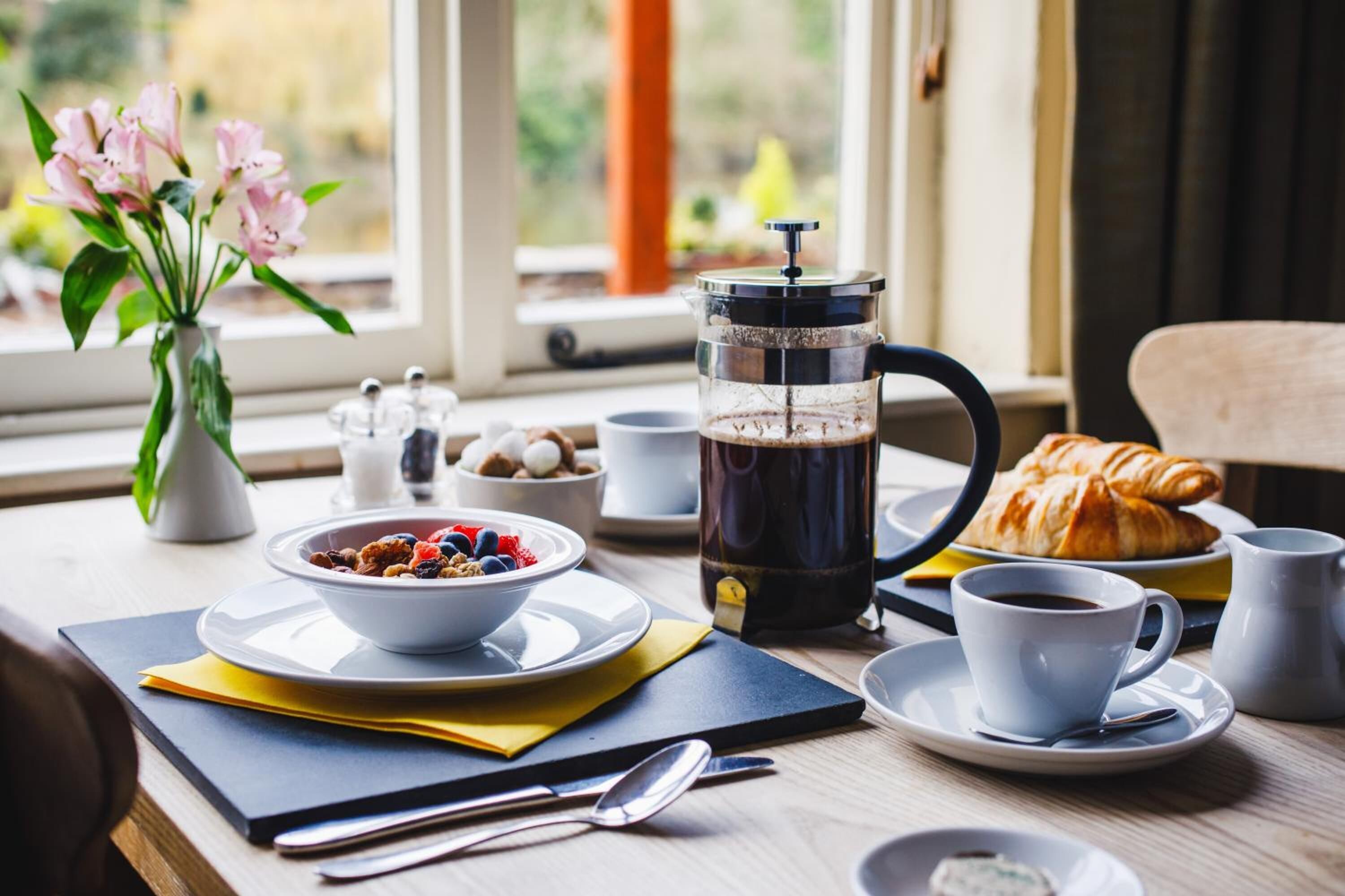 Una mesa de desayuno con cafetera, un croissant, un tazón de cereal y un florero.