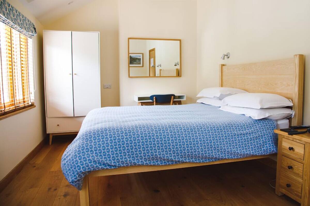 A bedroom with a bed, a wooden headboard, a bedside table with a phone, a desk with a chair, and a wardrobe.