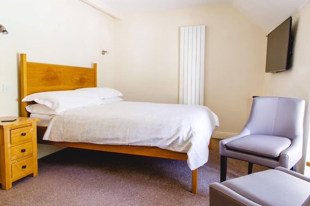 A bedroom with a wooden bed, a bedside table with drawers, a grey armchair, and a television mounted on the wall.