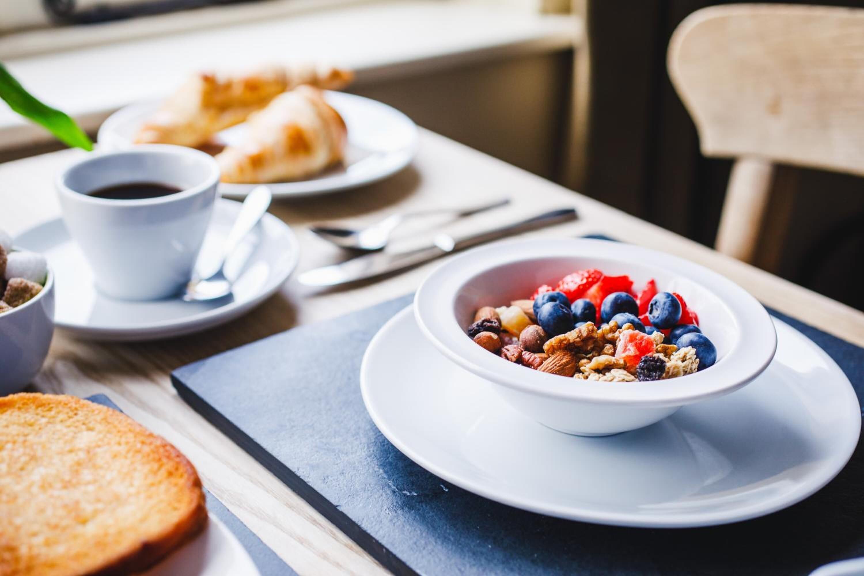 A breakfast setting with a bowl of cereal, a cup of coffee, and a croissant on a table.