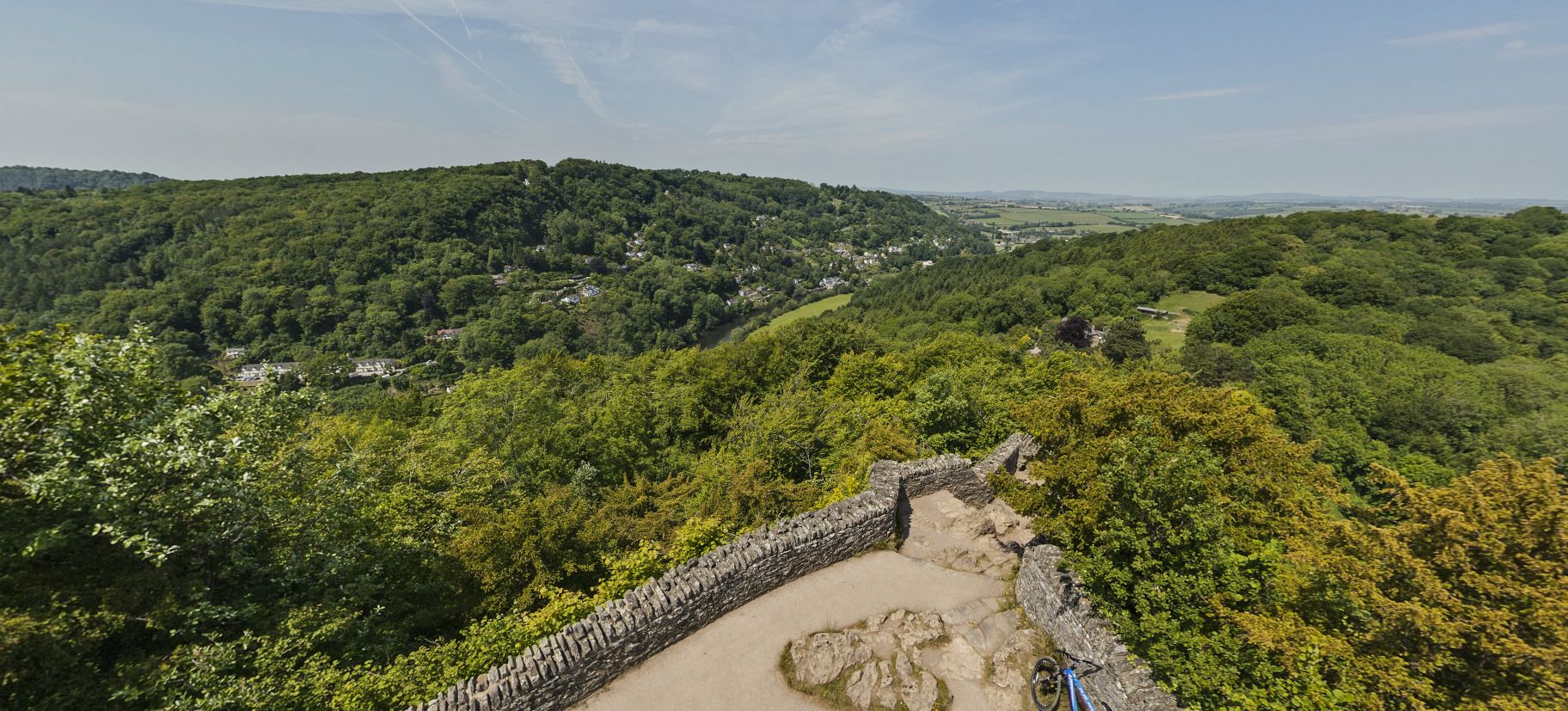 A stone pathway leading through a forested area with a view of a valley and distant buildings.