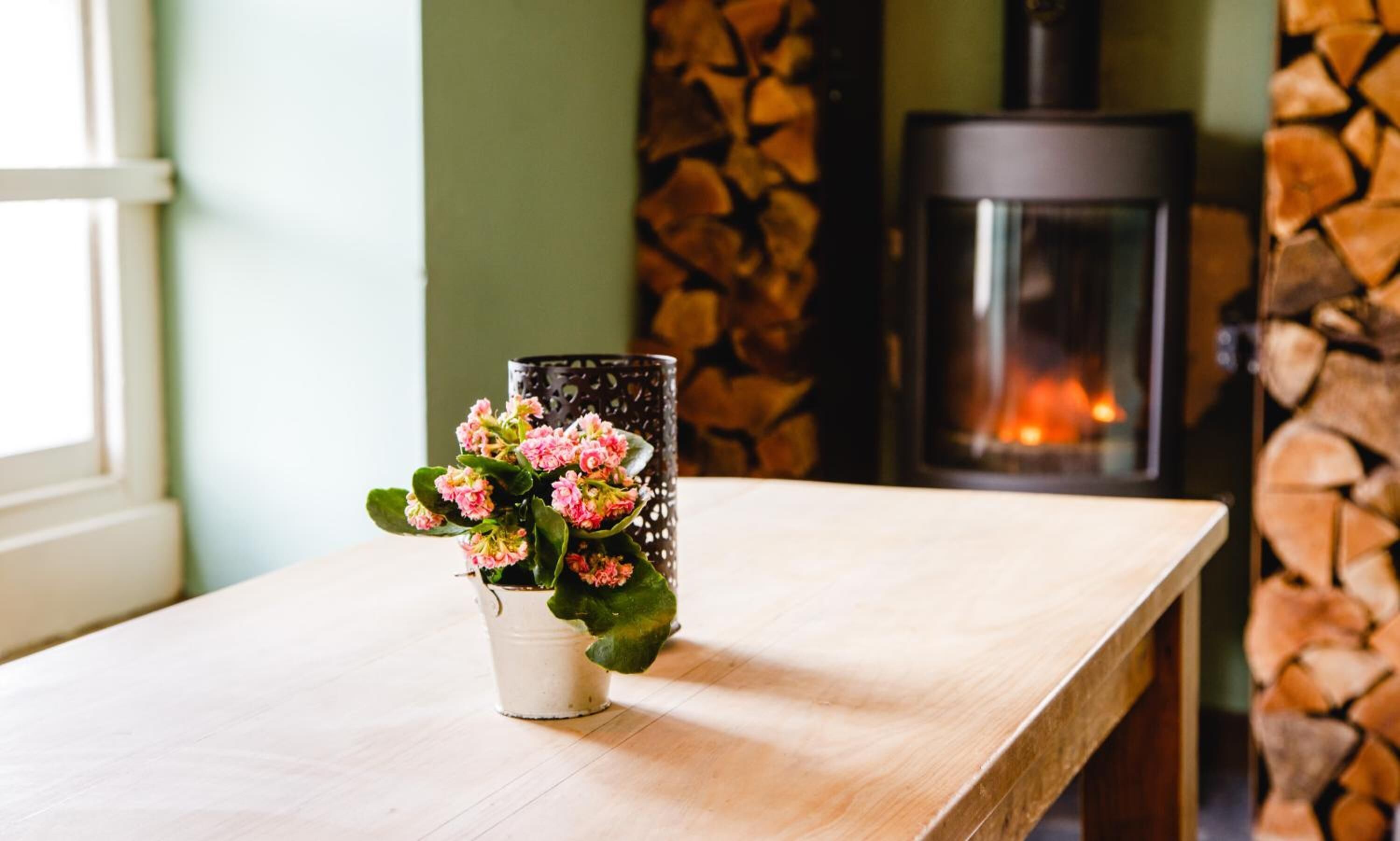 A wooden table with a white pot containing pink flowers. A black lantern with a patterned design is placed on the table. In the background, there is a stone wall with a fireplace.