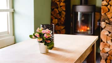 A wooden table with a white pot containing pink flowers. A black lantern with a patterned design is placed on the table. In the background, there is a stone wall with a fireplace.