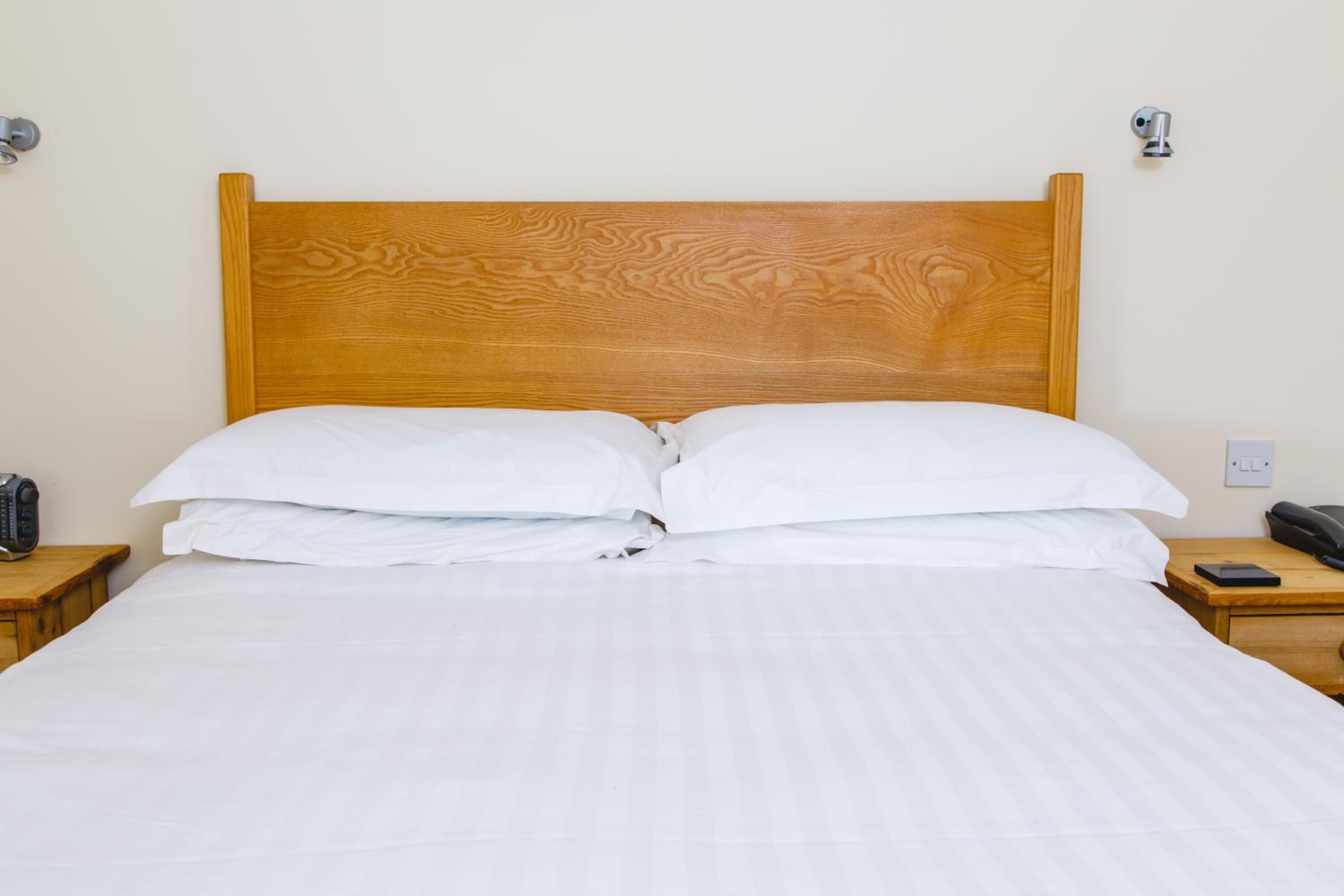 A neatly made bed with a wooden headboard, white bedding, and two bedside tables with a telephone and a power outlet on each.