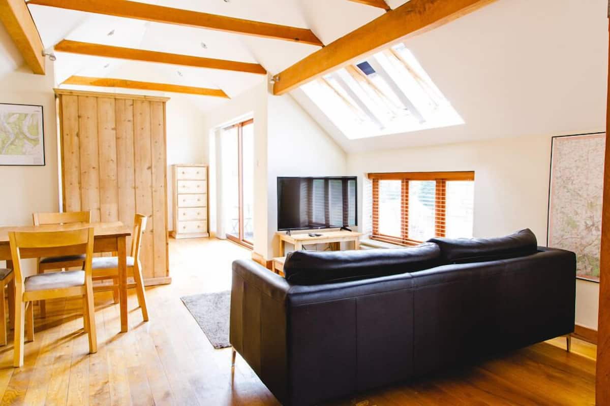 A spacious living room with a black sofa, wooden dining table, and chairs. A television is on a wooden stand, and there are large windows allowing natural light.