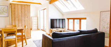 A spacious living room with a black sofa, wooden dining table, and chairs. A television is on a wooden stand, and there are large windows allowing natural light.