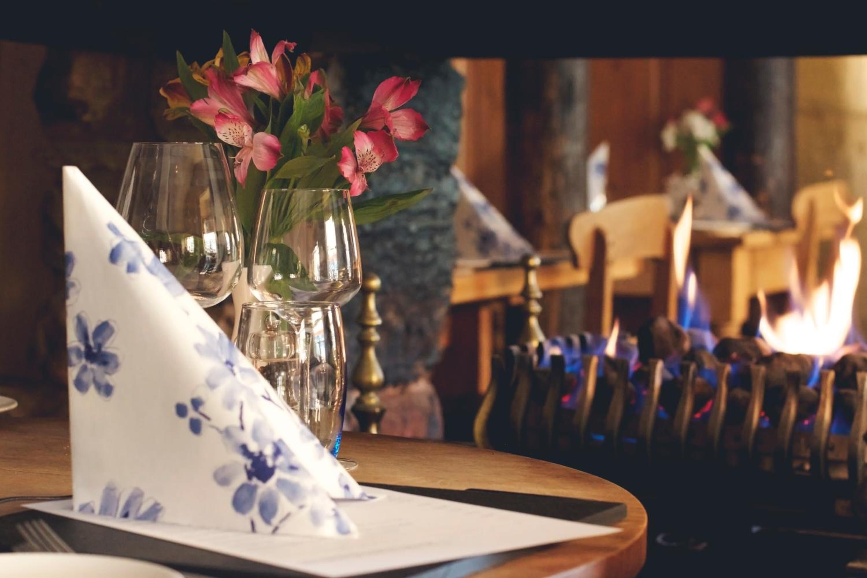 Une table à manger dressée avec une serviette à motifs floraux, deux verres à vin et un vase contenant des fleurs roses.