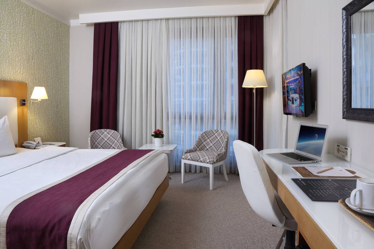 A hotel room with a large bed, a desk with a laptop, a chair, a small table with a lamp, and a TV on the wall.