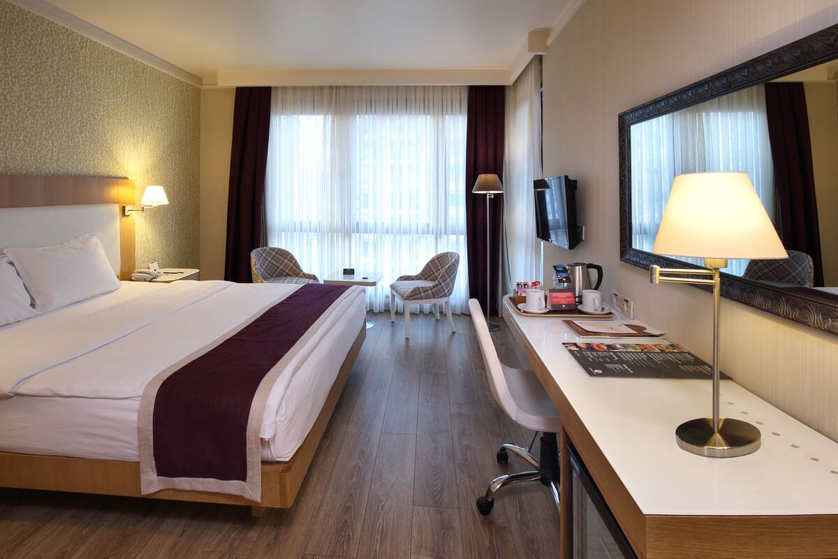 A hotel room with a large bed, a desk with a computer, a chair, a TV, and a window with curtains.