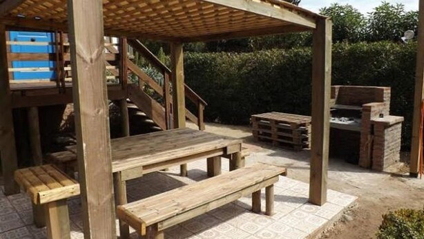 Uma área de estar ao ar livre com bancos de madeira, pérgola, churrasqueira e escadas que levam a um andar superior.