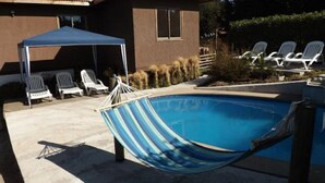 Uma piscina com uma rede, espreguiçadeiras e um espaço de estar coberto.