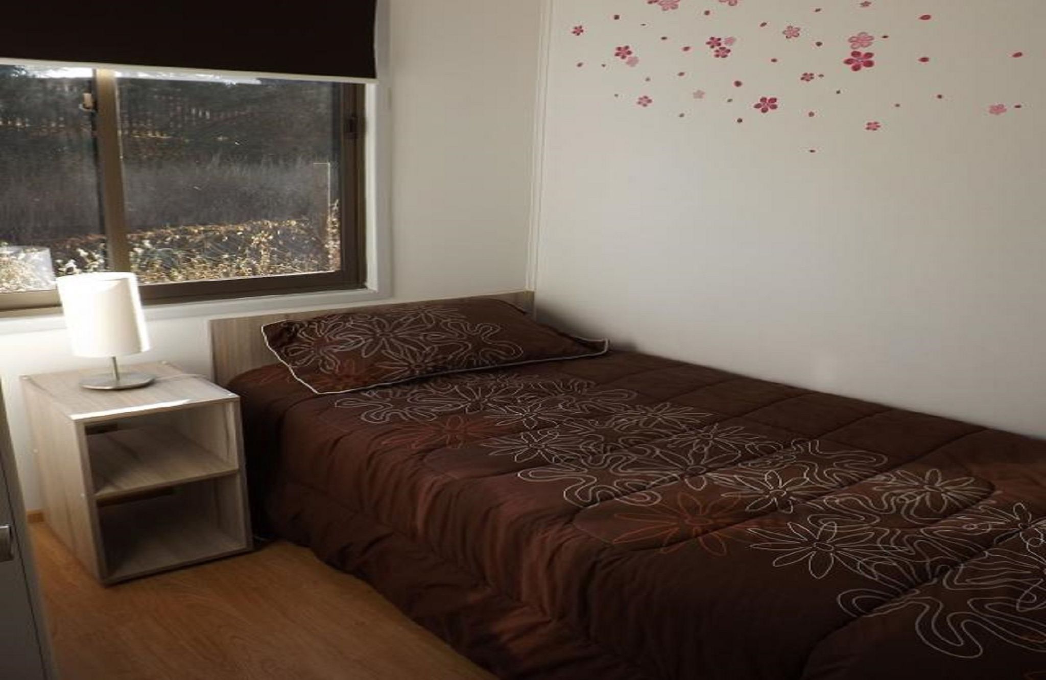 Una cama pequeña y bien hecha con un edredón marrón estampado con flores y una almohada a juego, una mesita de noche con una lámpara, y una ventana que ofrece vistas a árboles.