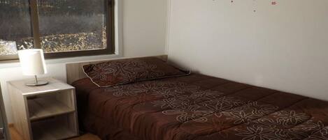 Una cama pequeña y bien hecha con un edredón marrón estampado con flores y una almohada a juego, una mesita de noche con una lámpara, y una ventana que ofrece vistas a árboles.